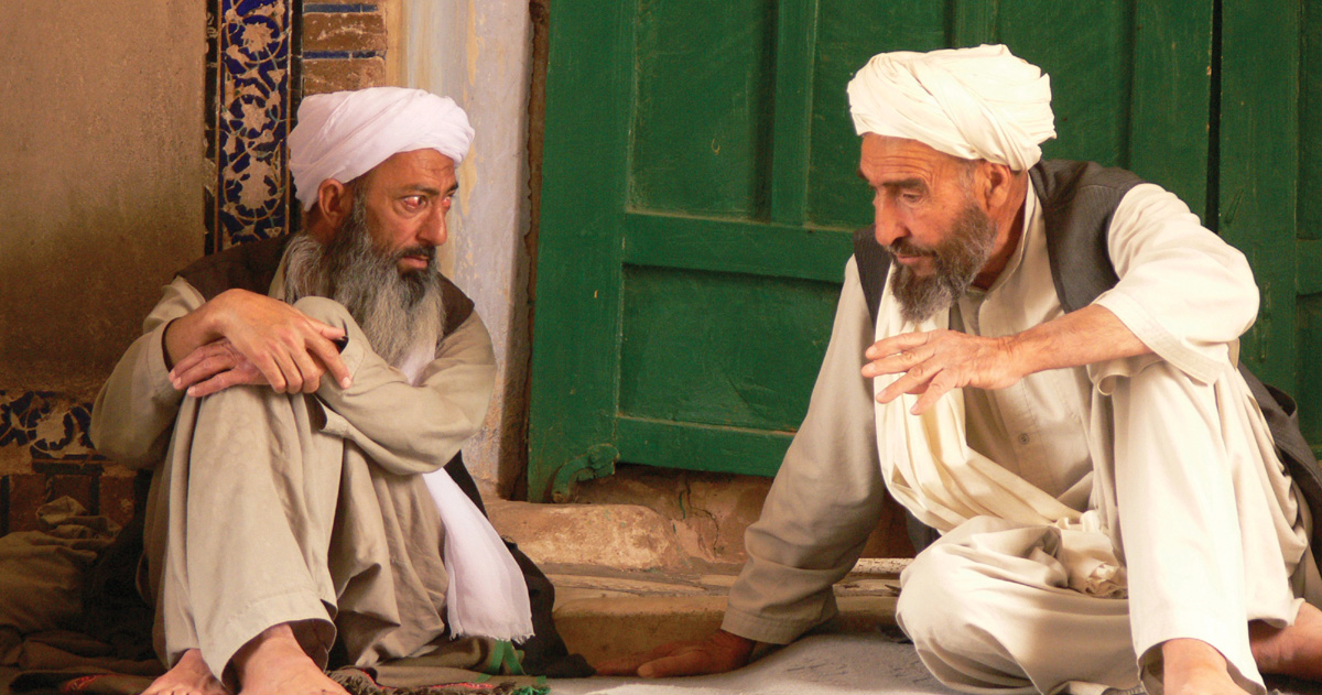 Two men are visiting while seated on the ground.