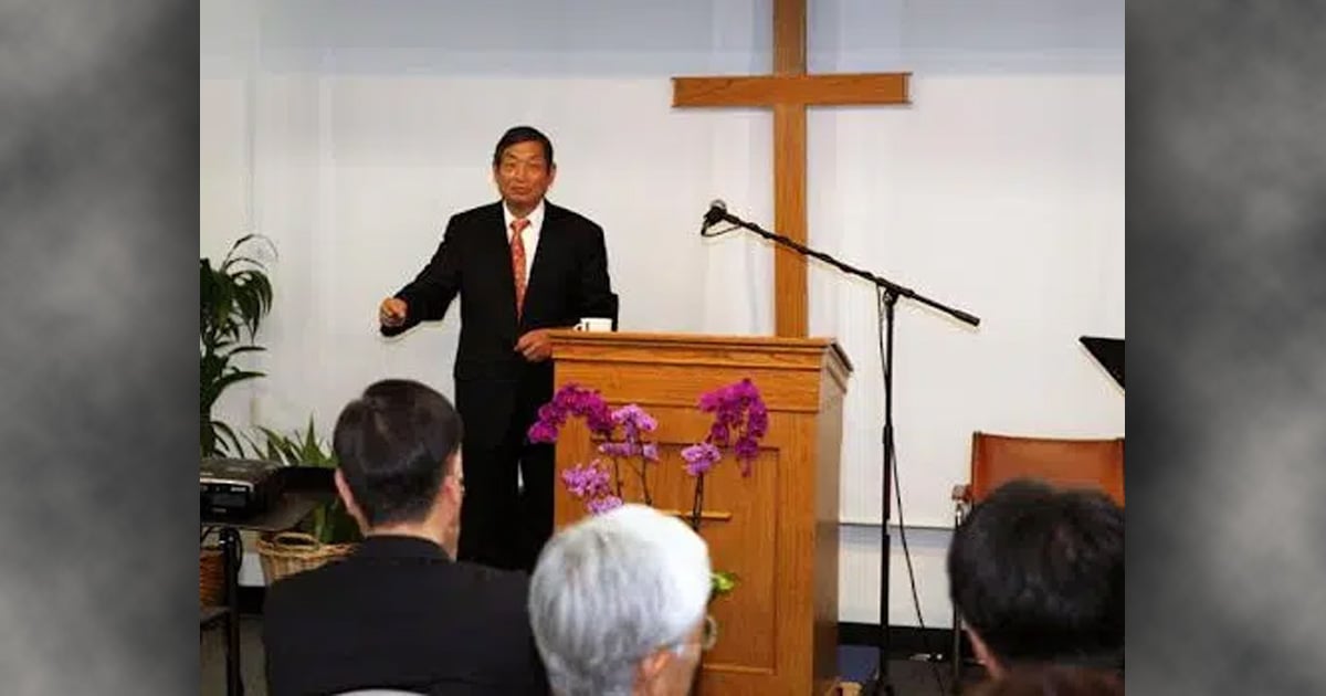 A smiling man is speaking behind a pulpit. There is a cross on the wall behind him.
