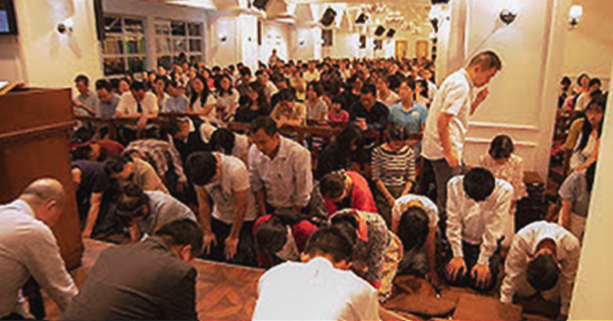 A building is filled with people praying. Several are on their knees with their heads bowed.