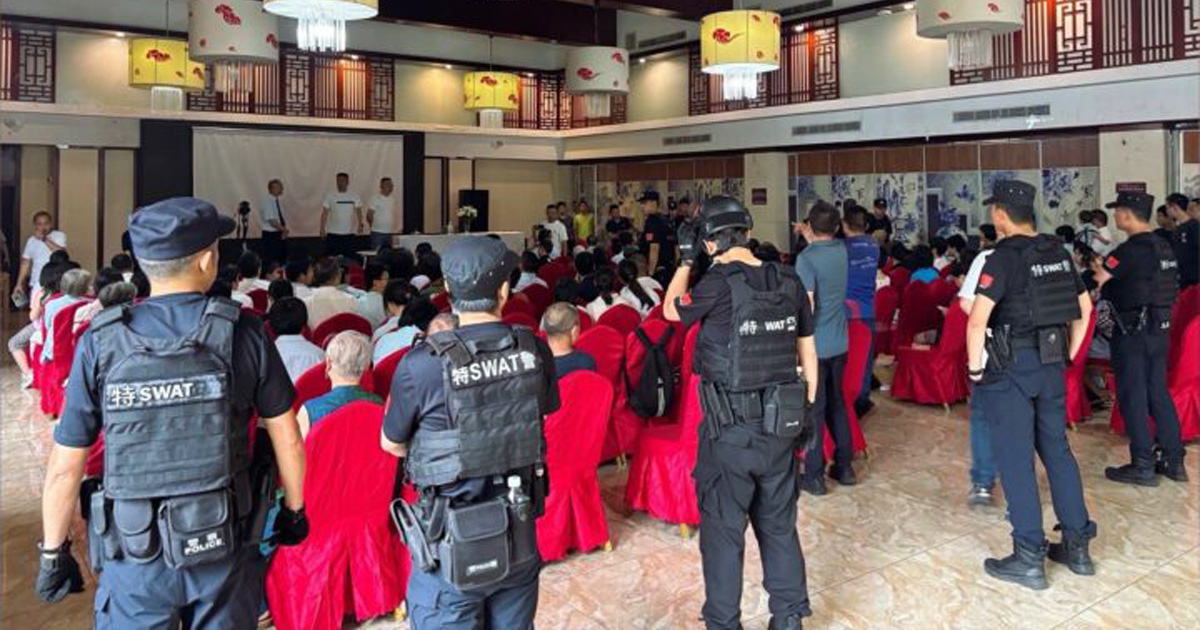 Officers are surrounding a group of people seated on chairs facing a stage. Officers are surrounding a group of people seated on chairs facing a stage.