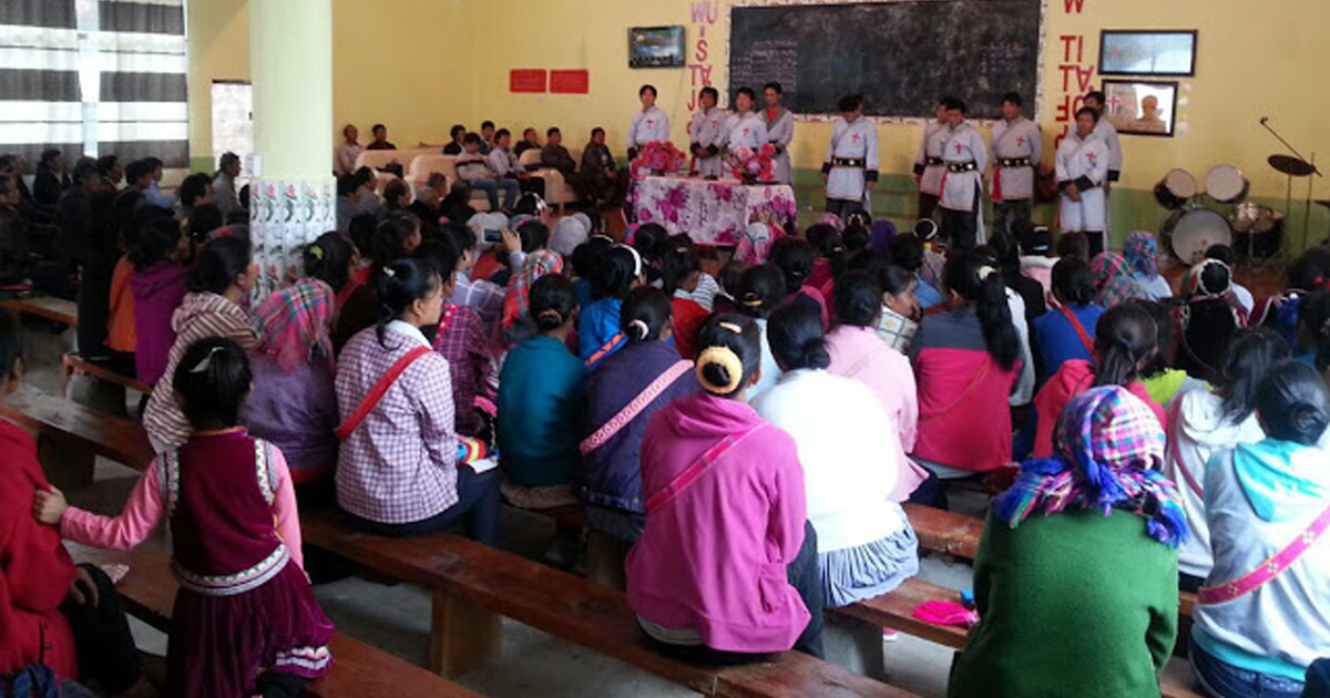 An audience is seated on wooden benches facing some individuals at the front of the meeting. An audience is seated on wooden benches facing some individuals at the front of the meeting.