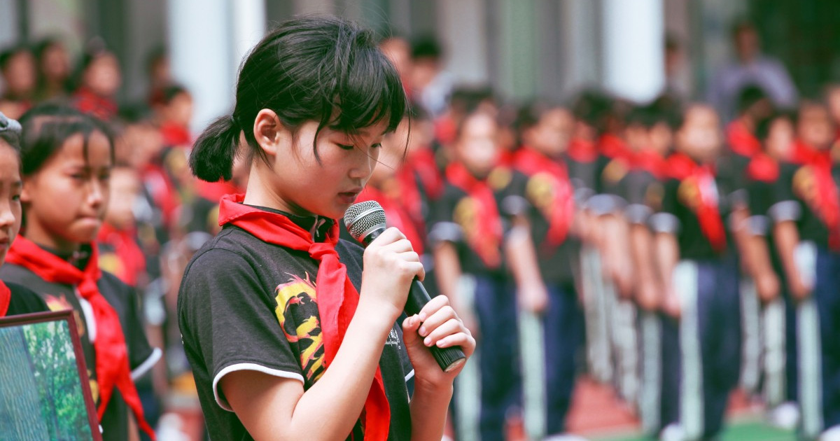 Photograph shows young girl with a microphone during an outdoor event. Children wearing red scarves stand in formation behind her on a green field, with spectators and buildings blurred in background. Photograph shows young girl with a microphone during an outdoor event. Children wearing red scarves stand in formation behind her on a green field, with spectators and buildings blurred in background.