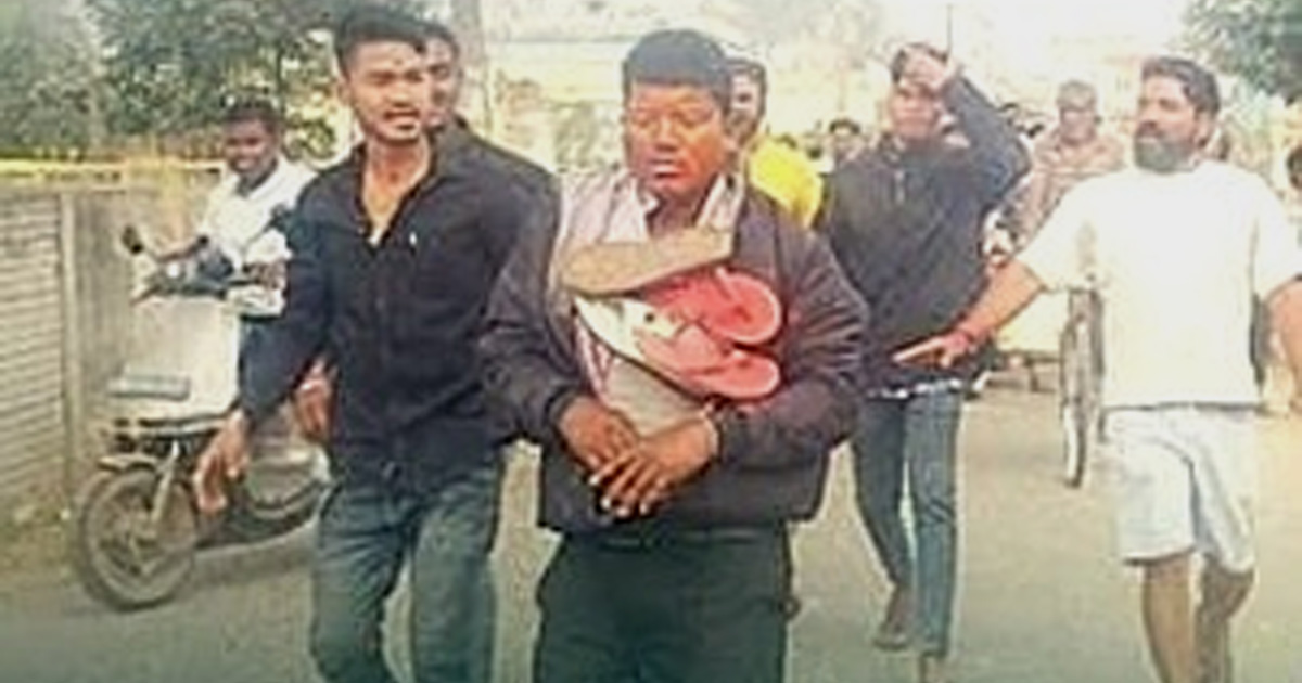 Pastor Bipin Bihari Naik appears to be stressed as he walks through the street with sandals draped around his neck. A crowd surrounds and is following him.