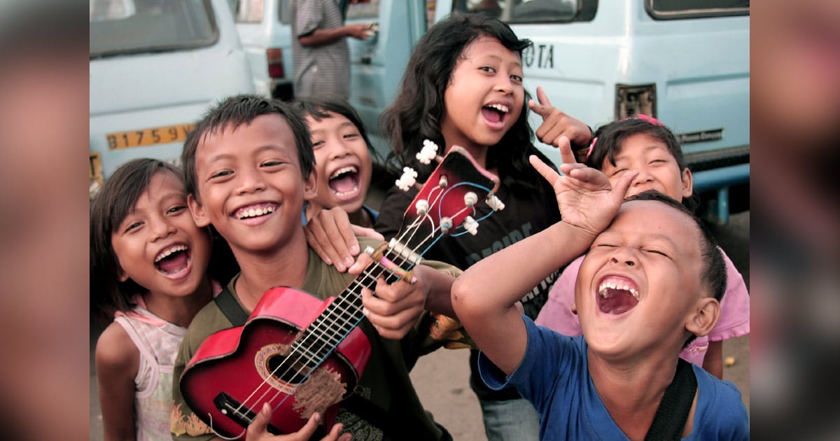 A group of children are smiling and laughing. One is playing an ukulele. A group of children are smiling and laughing. One is playing an ukulele.