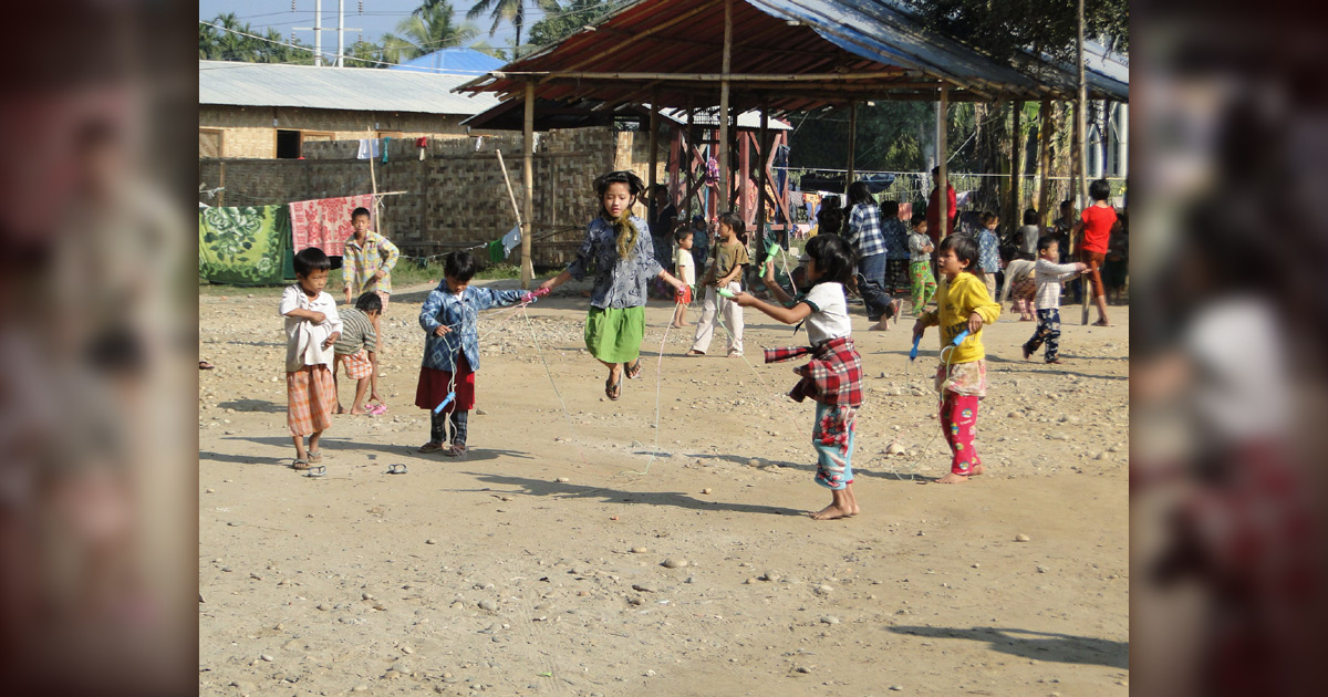 Children are jumping rope in the sunshine. There are other people in the background.