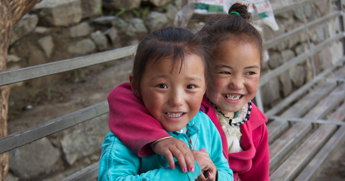 Two girls in brightly coloured clothing are hugging on an outdoor bench.