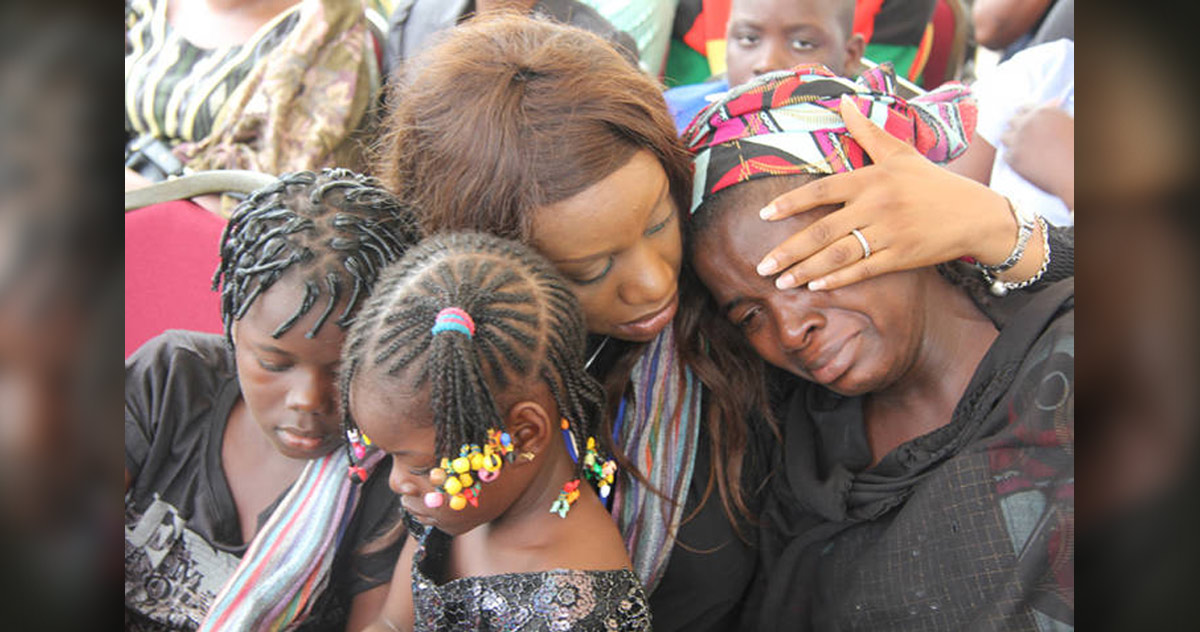 Photograph showing a woman comforting a tearful person wearing a colorful headscarf. Two children with braided hairstyles stand in the foreground, while adults in varied clothing fill background, conveying shared sorrow and support. Photograph showing a woman comforting a tearful person wearing a colorful headscarf. Two children with braided hairstyles stand in the foreground, while adults in varied clothing fill background, conveying shared sorrow and support.