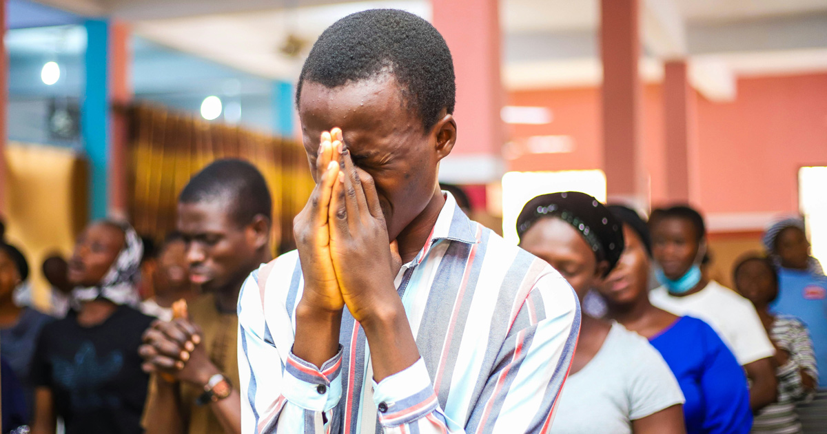 Several people are gathered together in prayer. The center of the image focuses on a man with his hands together and covering his face. He is focused on prayer. Several people are gathered together in prayer. The center of the image focuses on a man with his hands together and covering his face. He is focused on prayer.