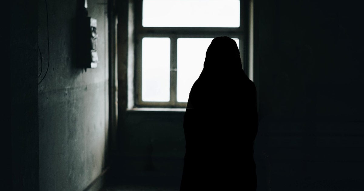 The silhouette of a woman in a head covering can be seen against a window. The room around her is very dark except for the daylight coming from the window.