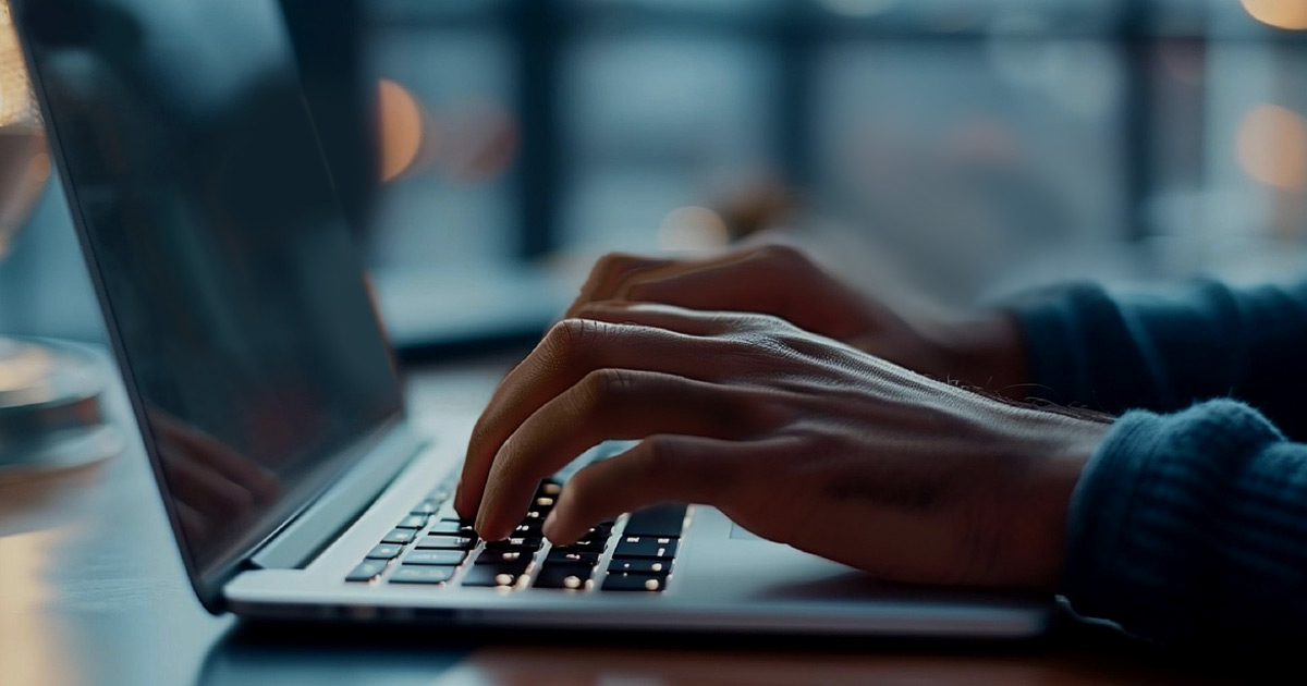 A man's hands are shown typing on a laptop in a dimly lit room. A man's hands are shown typing on a laptop in a dimly lit room.
