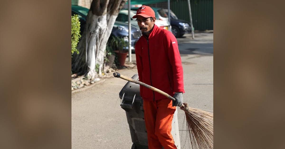 A man dressed in orange overalls and cap is holding a straw broom and pulling a garbage bin.