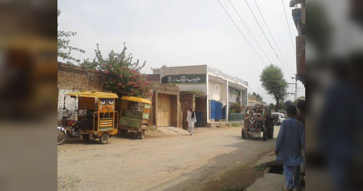Photograph shows a village street with small shops, roadside vendors, and local traffic and a few pedestrians. Photograph shows a village street with small shops, roadside vendors, and local traffic and a few pedestrians.