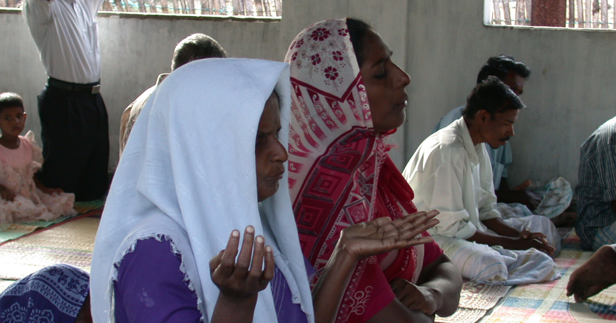 Christians are seated on floor mats as they pray together. Two women are in the foreground.