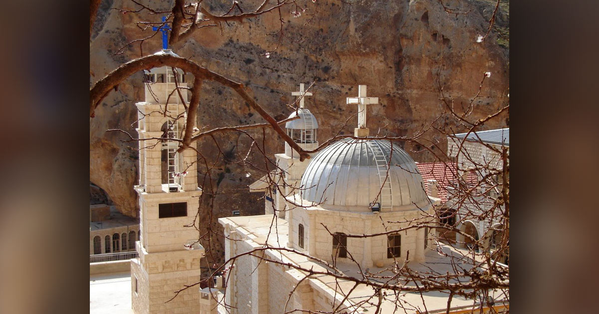 Photograph of the St. Teqla Church complex built against a rugged rock cliff, viewed through leafless branches. White domes, multiple crosses, bell tower, arched windows, and weathered masonry highlight religious architecture.