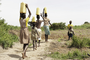 Children in Uganda; girls walking down a road with water jugs on their heads with little boys watching - Photo: Pixabay / Numbercfoto