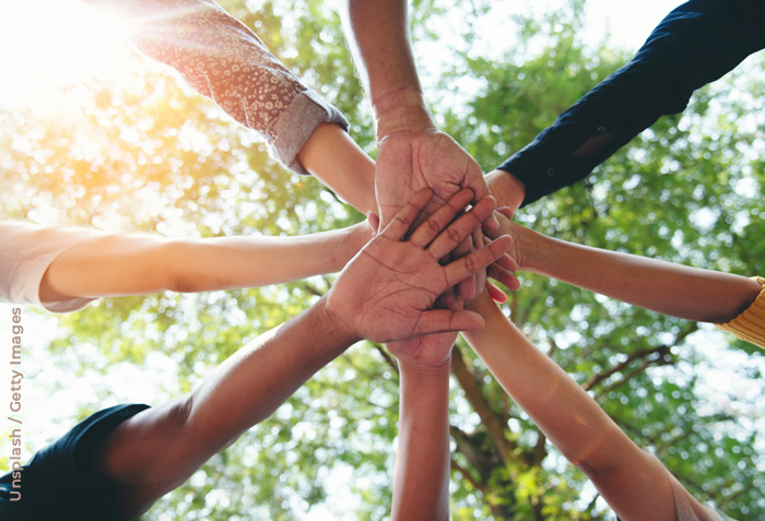 A group of hands are seen one on top of another against the sunlight bursting through tree brances.