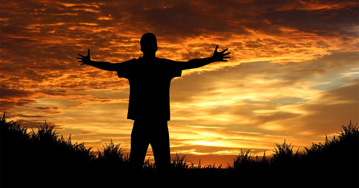 A man is standing with his arms raised. A sunset in the background creates a silhouette of him and the surrounding grass.