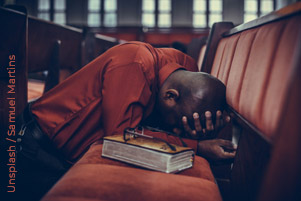 A man is kneeling at a church bench, praying fervently. His glasses rest on the Bible beside him.