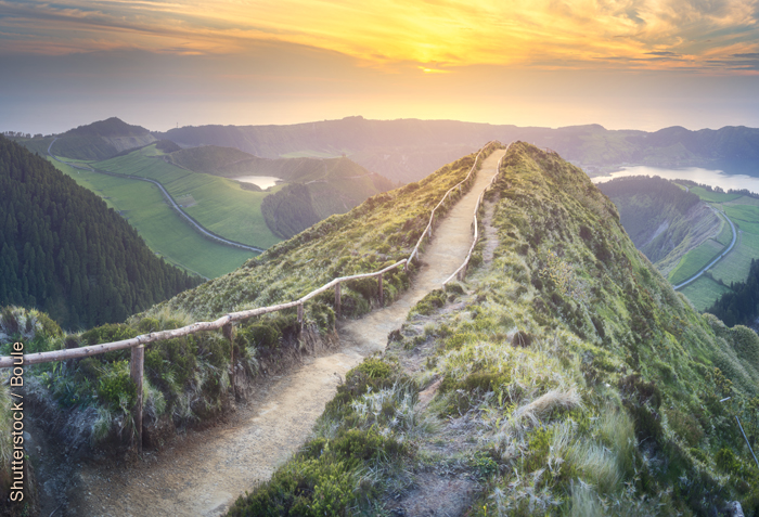 A mountain path follows the ridge of a mountain and the sun sits just above the horizon.