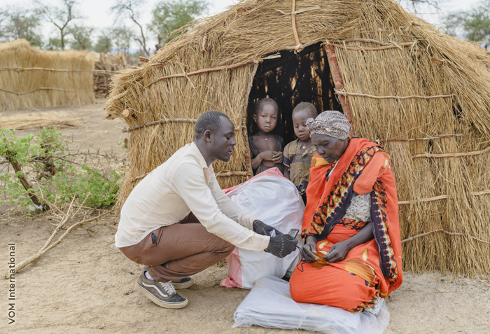 A man is showing something to a woman who is just outside a small straw hut. There are two children in the doorway.